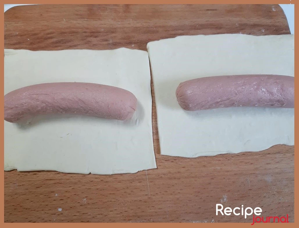 Рабочую поверхность припудриваем небольшим количеством муки, размороженное слоёное тесто слегка раскатываем и разрезаем на прямоугольники по размеру, чтобы поместилась сосиска. В Центр прямоугольника кладем сосиску.